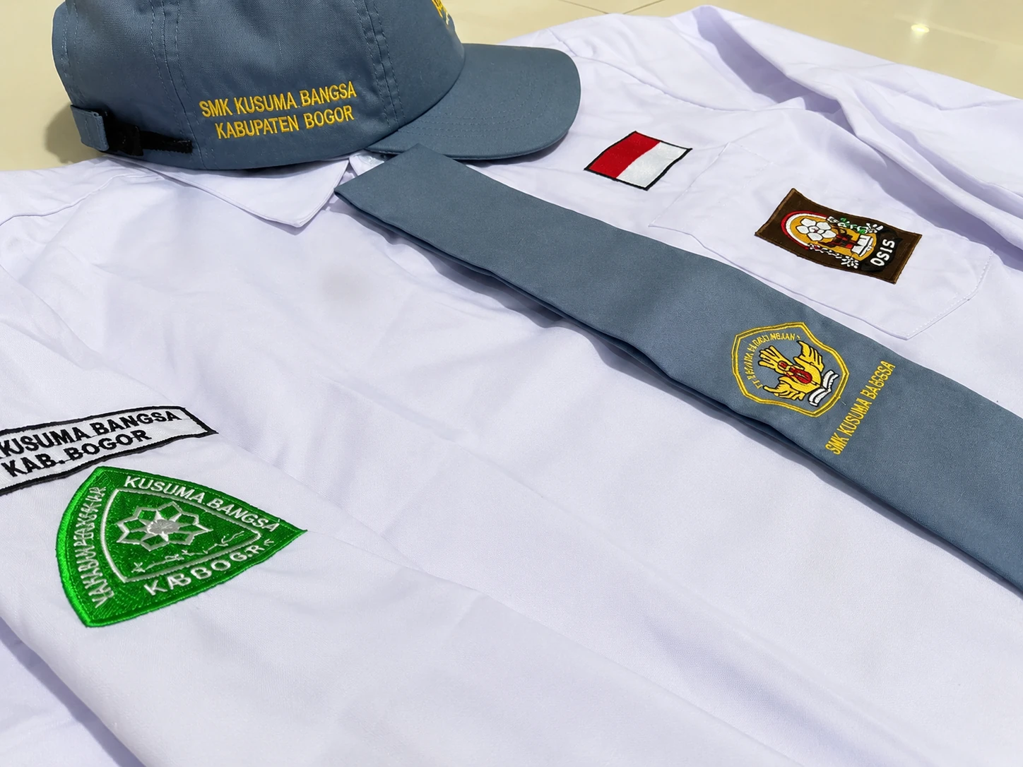 Seragam putih sekolah SMK Kusuma Bangsa dengan badge, topi abu-abu, dan dasi produksi JAP Solusindo